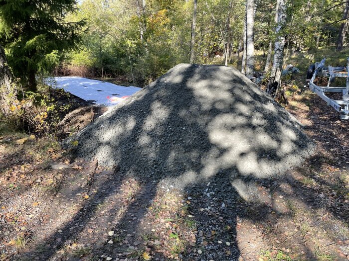 En hög med grus ligger på marken i en skogsmiljö, omgiven av träd och löv, med ett vitt skyddande täcke i bakgrunden.