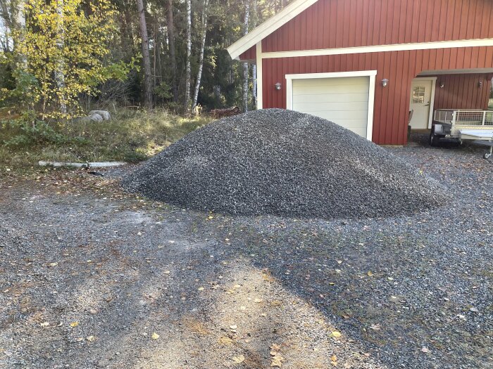 En stor hög med grus ligger bredvid ett rött hus med garageport, omgiven av träd och natur.