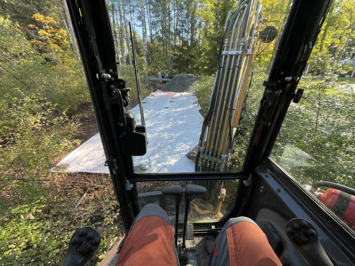 Vy från grävmaskin i skogen med grusplan och vita presenningar, inifrån förarhytten med körspakar och grävmaskinsarm.