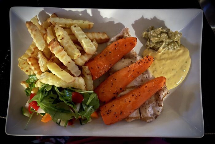 Tallrik med grillad kyckling, pommes frites, ugnsgrillade morötter, sallad och guacamole med bearnaisesås.