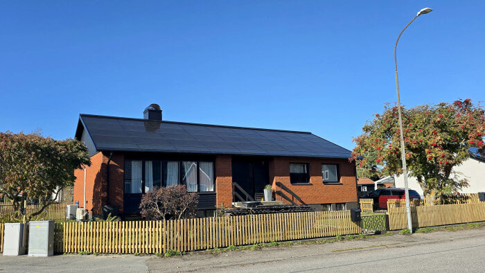 Rött tegelhus med svart soltak och staket framför, blå himmel och korsande gata i förgrunden.