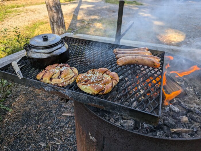 Ramslökskorv och kanelbullar grillas över öppen eld på ett galler, bredvid en svart kastrull.