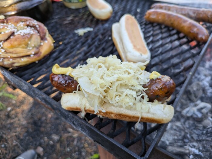 Ramslökskorv i bröd med senap och surkål på grillgaller, kanelbulle och korvbröd vid sidan.