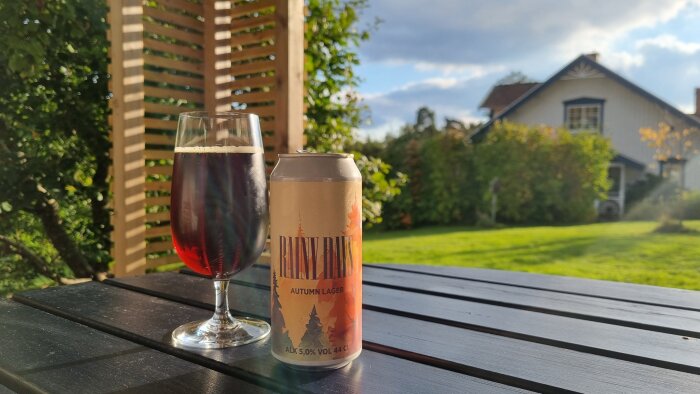 Ett glas med mörk öl och en burk märkta "Rainy Days Autumn Lager" står på ett bord i trädgården, med ett hus i bakgrunden.