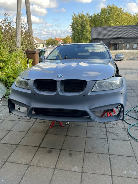 En grå BMW-bil med nyligen monterad front, uppställd på stödjack på en uppfart. Bakgrund med träd och hus.