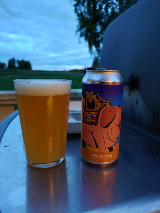 Ett glas hazy IPA-öl bredvid en färgglad ölburk med design, placerat på en grill utomhus med en bakgrund av äng och kvällshimmel.