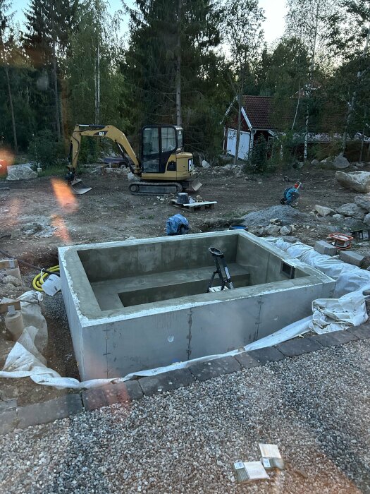 Betongpool under byggnation med en grävmaskin och verktyg i bakgrunden, omgiven av träd och ett rött hus.