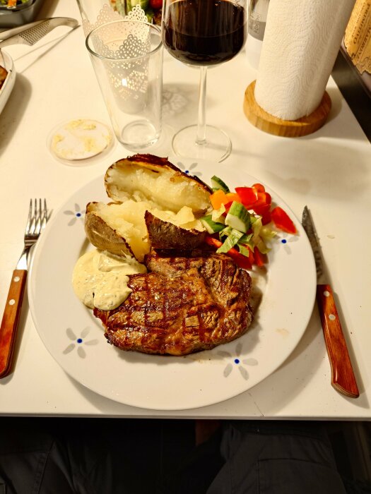 Grillad entrecote och biff med bakad potatis och grönsaker på en tallrik, glas vin i bakgrunden.