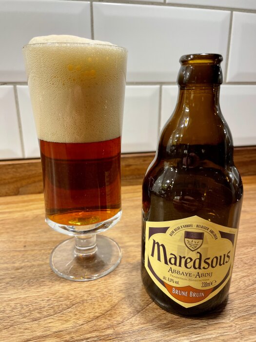 Brunt belgiskt öl Maredsous i glas och flaska på träbord, skummande dryck mot vit kakelvägg.