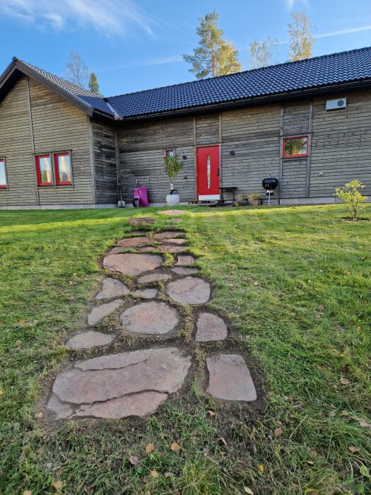 Stenlagd gång av oregelbundna stenar med gräs mellan, leder till en modern, träklädd villa med röd dörr och svarta takpannor.