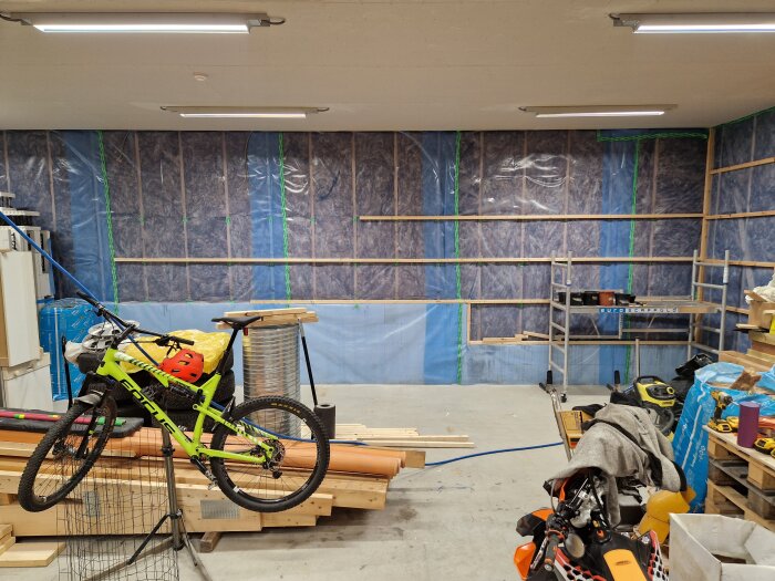 Stort garage under renovering med isolerade väggar, byggmaterial utspritt och en grön mountainbike upphängd framför arbetsplatsen.