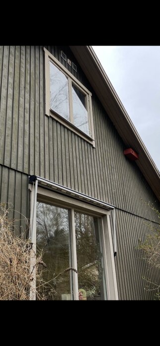 Svart träfasad av ett hus med två fönster, varav ett övervåningsfönster och ett under, där markisen nyligen tagits bort.