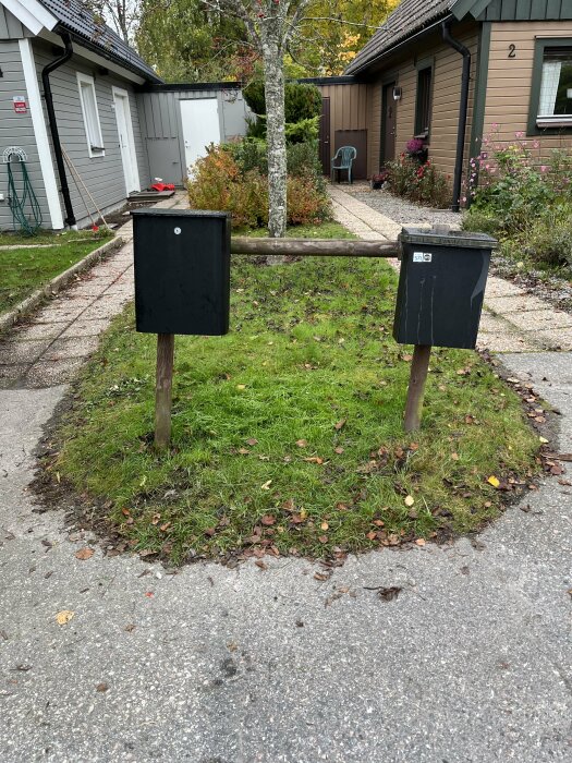 Två brevlådor monterade på träreglar över en gräsyta framför två hus, omgivna av buskar och träd.