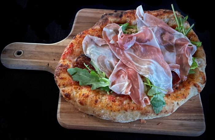 Pizza med tomatsås, mozzarella, serranoskinka och ruccola på en träskärbräda.