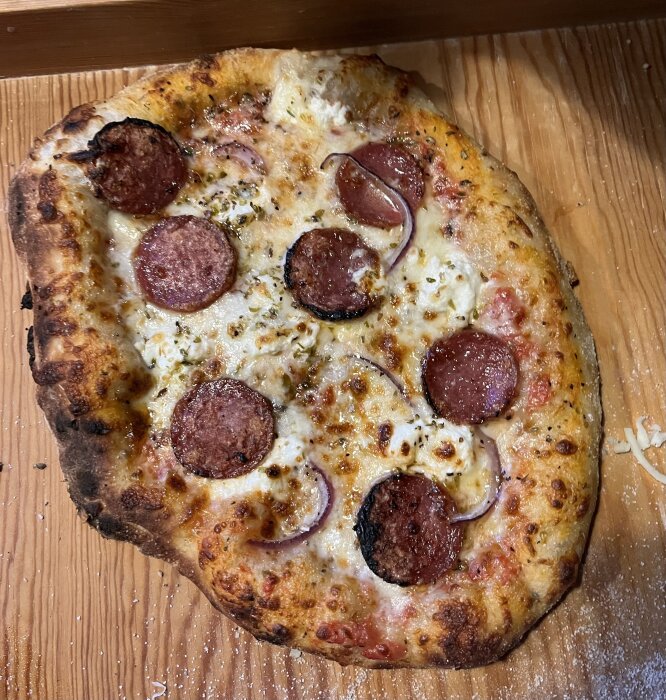 Hemgjord pizza med tomatsås, mozzarella, salami och rödlök på träbricka. Något bränd kant, gyllenbrun ost, använts under pizzakväll.