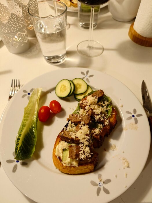 Bookmaker toast med kött, gurkskivor och sallad på en tallrik, serverad med vin och vatten i glas.