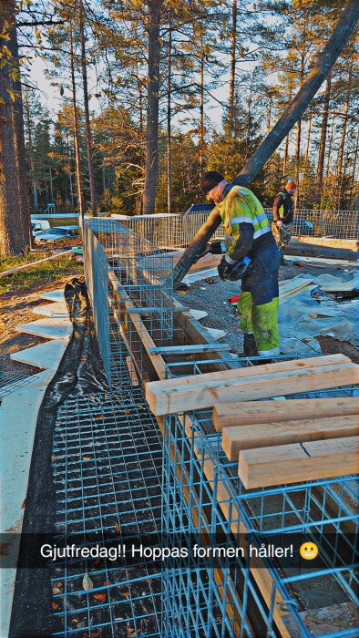 Person gjuter kantbalk i träform med betongpump i skogsmiljö. Byggprojekt med armeringsjärn och byggmaterial runtomkring.