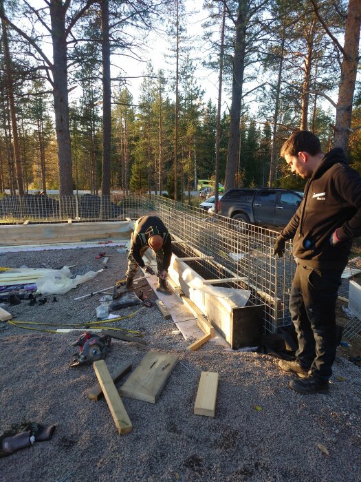 Två personer arbetar utomhus med en kantbalksform; omgivna av tallar och byggmaterial, inklusive plankor och verktyg. En bil syns i bakgrunden.