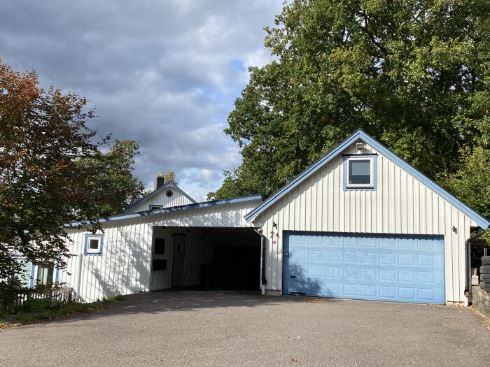 Vit villa med blå garageport och carport, omgiven av träd. Asfalterad uppfart, molnig himmel syns i bakgrunden.