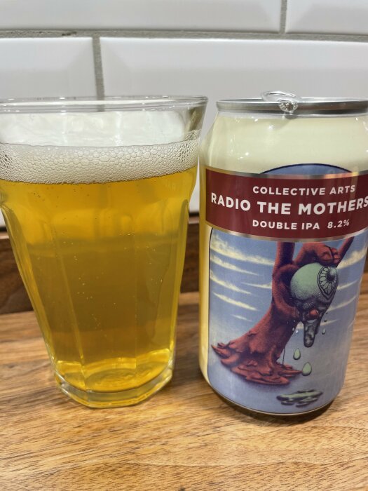 Ett glas med gyllene öl bredvid en burk med etiketten "Collective Arts Radio The Mothers Double IPA 8.2%", placerade på en träyta.