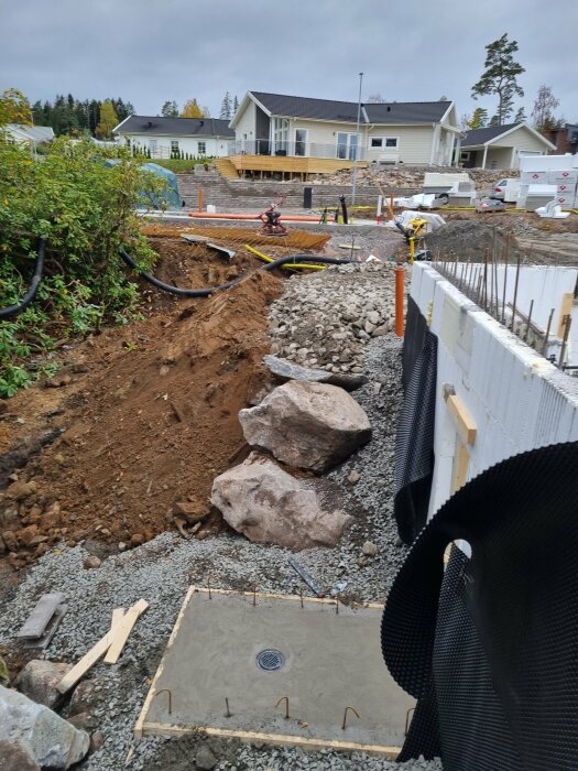 Betongplatta med brunn vid källaringång på en byggarbetsplats, omgiven av jord och stora stenar, med hus i bakgrunden.