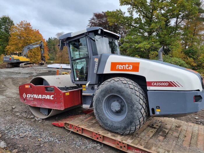 Vältmaskin Dynapac CA3500D på en byggarbetsplats, omgiven av grus och en grävmaskin i bakgrunden, höstlöv på träden.