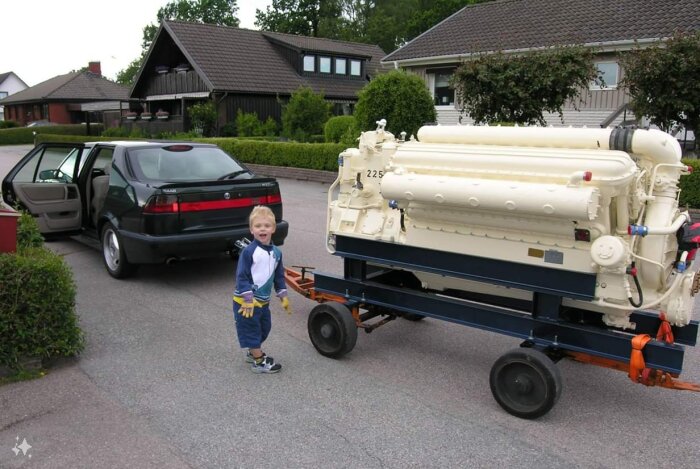 Barn bredvid en stor motor, Zvezda M50F6L, på vagn framför en bil i ett bostadsområde.