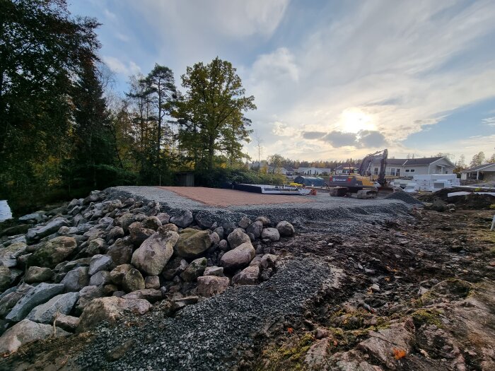 Byggarbetsplats med grus och sten, omgiven av träd och hus, en grävmaskin står nära solnedgången i bakgrunden.