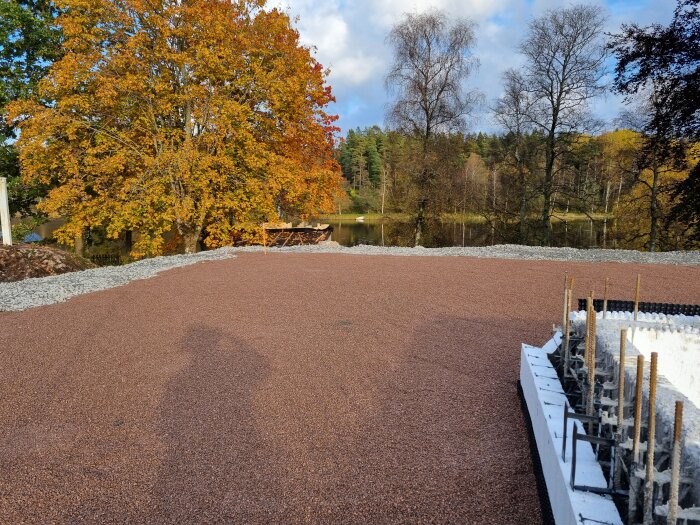 Material förberett för byggprojekt med utlagt makadam och krattad flis, omgiven av träd med höstlöv vid en sjö.