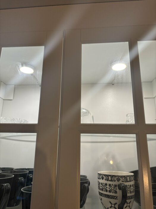 Köksskåp med glasdörrar och två LED-spotlights; en fungerar, en är släckt. Svartvit kopp och glas inuti skåpet.