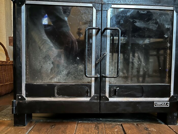Svart glaslucka på en kamin med texten "IMPULS" i nedre hörnet. Reflektion av en person på glaset.