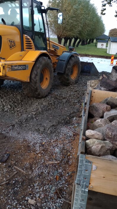 En gul traktor med skopa kör över grus och sten på en byggarbetsplats. I förgrunden syns en låda fylld med stora stenar.