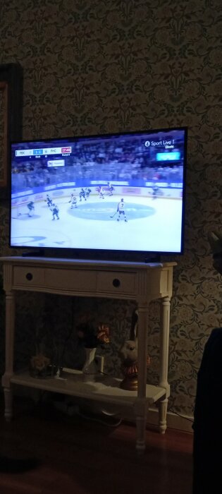 TV visar en hockeymatch mellan FBK och FHC i period 2. TV:n står på en vit bänk med blommig tapet i bakgrunden.