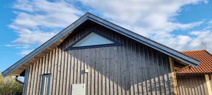 Fritidshus med träpanel på norrfasaden och triangulärt fönster, målat med Falu svart slamfärg under en klarblå himmel.