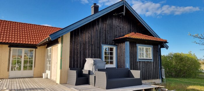 Fritidshus med ny svartmålad panel och rött tak, möblerad altan i förgrunden och grönska i bakgrunden under klar himmel.