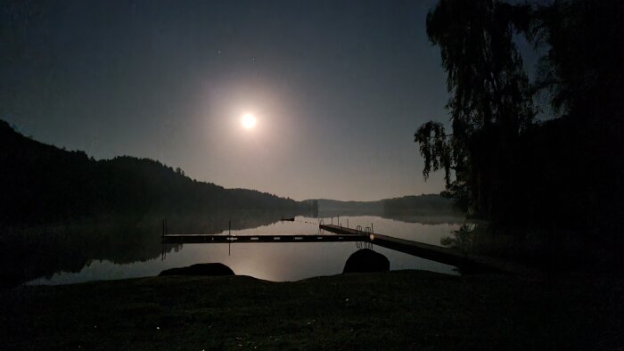 Månsken över en sjö, med brygga och siluetter av träd och kullar. Vattnet är lugnt och reflekterar det ljusa månskenet.