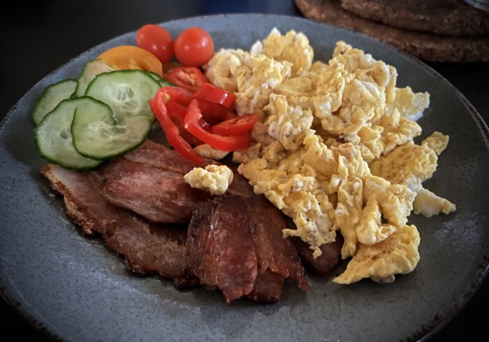 Tallerken med stekfläsk, äggröra, gurkskivor, paprika och cocktailtomater; del av en lyxig hemmabrunch.