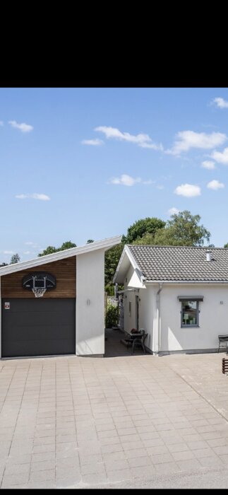 En modern villa med putsad fasad, garage och basketkorg på garageporten, med trädgård och blå himmel i bakgrunden.