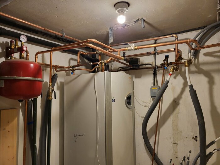 Värmepumpsystem med isolerade rör och kopparrör i en källare, kopplat till rör och tankar för golvvärme och ventilation.