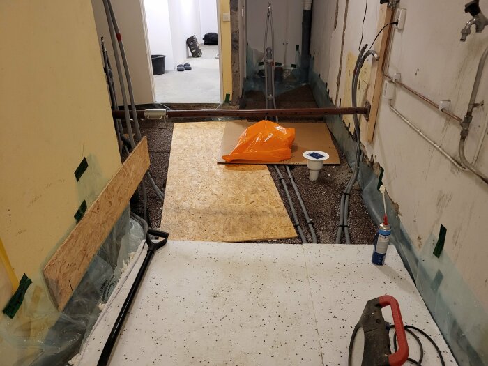 Rörinstallation i renovering: grå rör längs väggen, orange säck, skiva på golvet, betongplattor och verktyg i rummet; höger sida visar väggdetaljer.