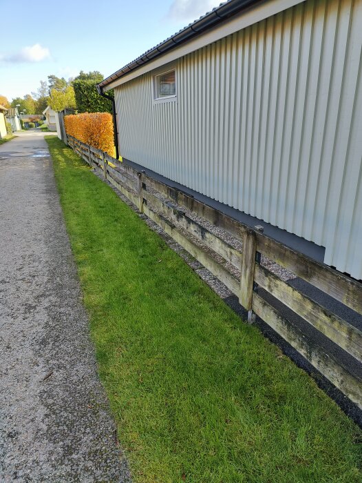 Slitet trästaket med lavapåväxt längs en grön gräsmatta och gångväg vid ett hus med vit fasad.