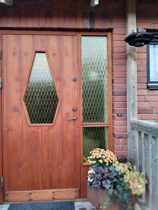 Entrédörr i trä med glasdetaljer, omgiven av tegelväggar och ny fasadbelysning med liten glasglob, samt blomplantering vid sidan av dörren.