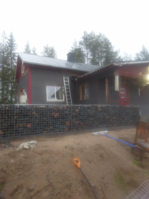 Grått hus under renovering med stege lutad mot väggen, stenfyllda gabioner och verktyg på marken i en skogsmiljö.