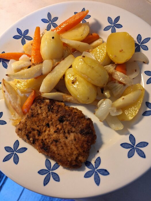 Ugnsrostade rotfrukter med potatis, morötter, palsternacka och lök, serverad med hemgjord schnitzel på fläskytterfilé på en tallrik med blå blommor.