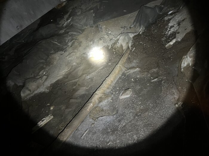 Bild av en mörk betongyta med en vattenpöl och ett avloppsrör, belyst av en ficklampa. Ytan i ett kryputrymme ser sliten ut.