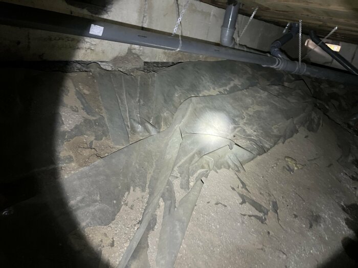 Bilden visar en pöl under ett hus, omgiven av plast och rör. Det verkar vara en undergolvsinstallation.