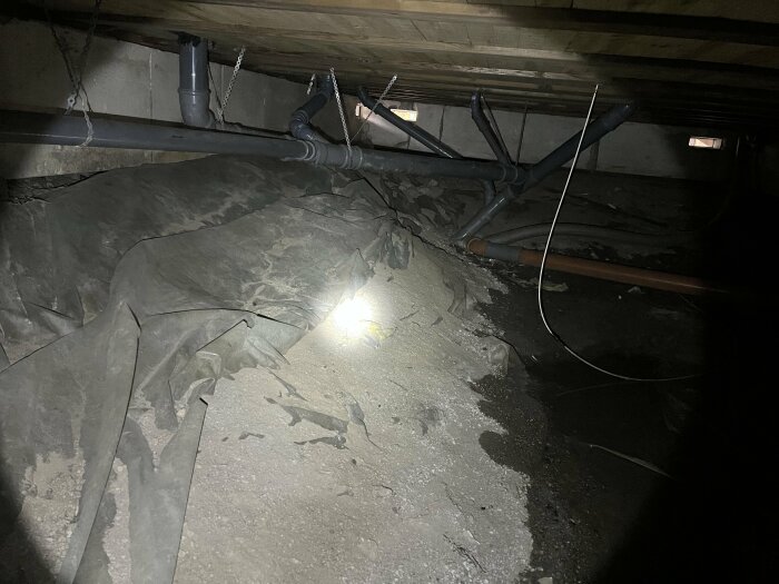 Vattenpöl på marken under husgrund med rör och plastskydd synliga, svag belysning.