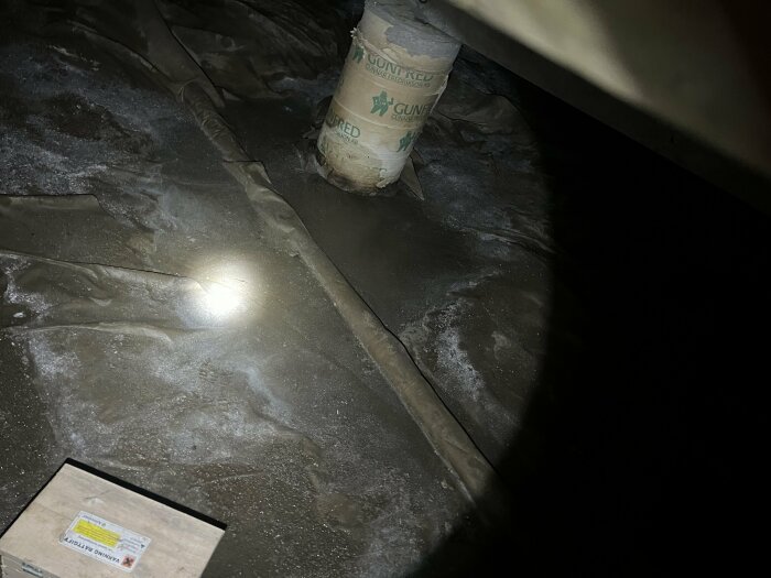 Vattenpöl på golvet i en krypgrund, omgiven av plastfolie och ett rör, med skugga från en ficklampa.