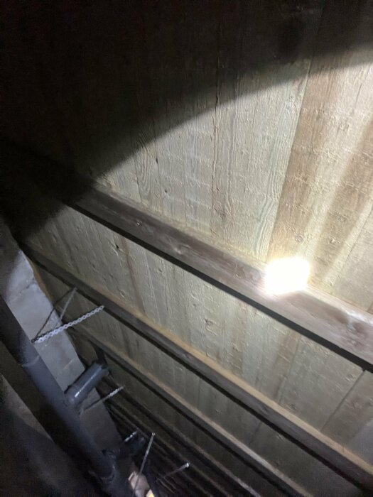 Träkonstruktion i källare med belysning, synliga rör och kedjor hängande från taket, del av en diskussion om vattenpöl och dränering.