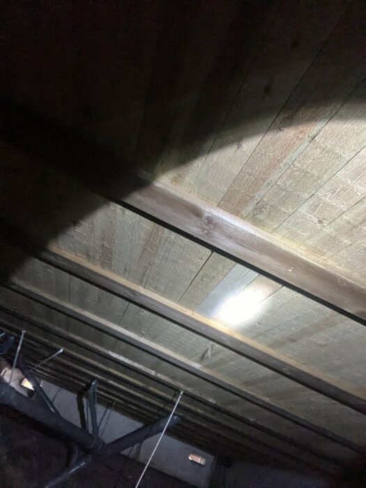 Trätak med skugga och belysning, synliga bjälkar och en cementvägg i bakgrunden, relaterat till frågeställning om vattenpöl i källaren.